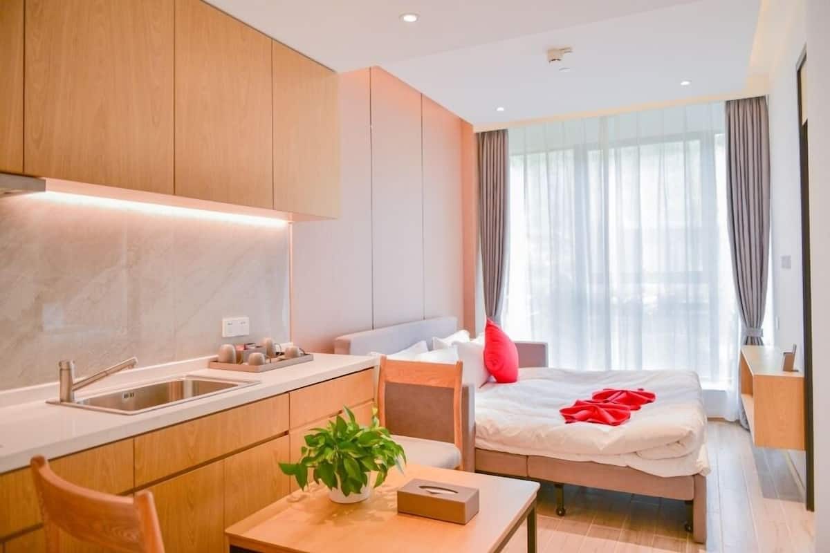 Ein modernes Hotelzimmer mit Küchenzeile, einem Bett mit rotem Kissen und einem großen Fenster mit Vorhängen.