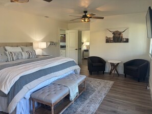 Une chambre à coucher avec un grand lit, deux chaises, un banc, un ventilateur de plafond et une porte donnant sur une autre pièce.