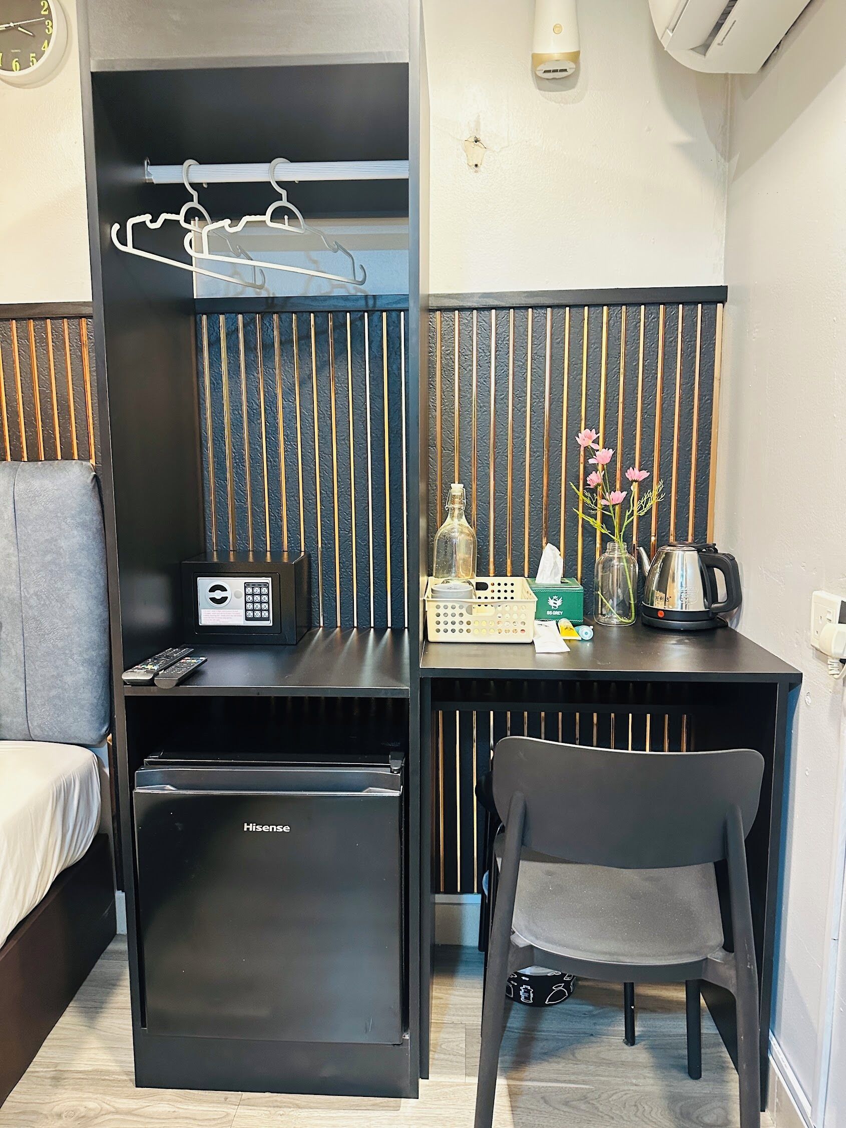 Ein kompaktes Hotelzimmer mit einem Bett, einem kleinen schwarzen Kühlschrank, einem Schreibtisch mit Wasserkocher, einem Sessel und einer Wand-Klimaanlage.