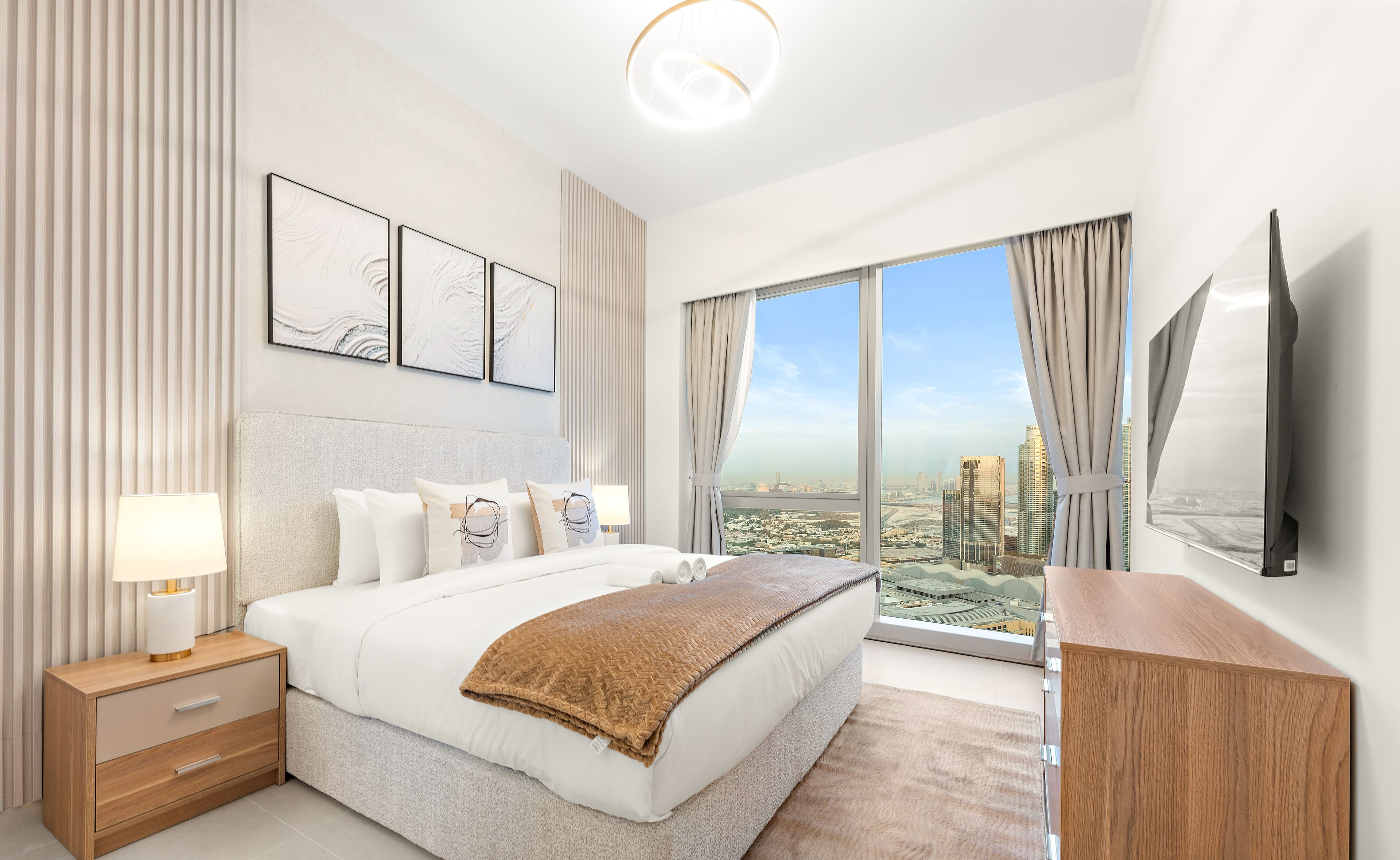 A modern hotel room with a large bed, a bedside table with a lamp, a flat-screen TV mounted on the wall, and a large window offering a city view.