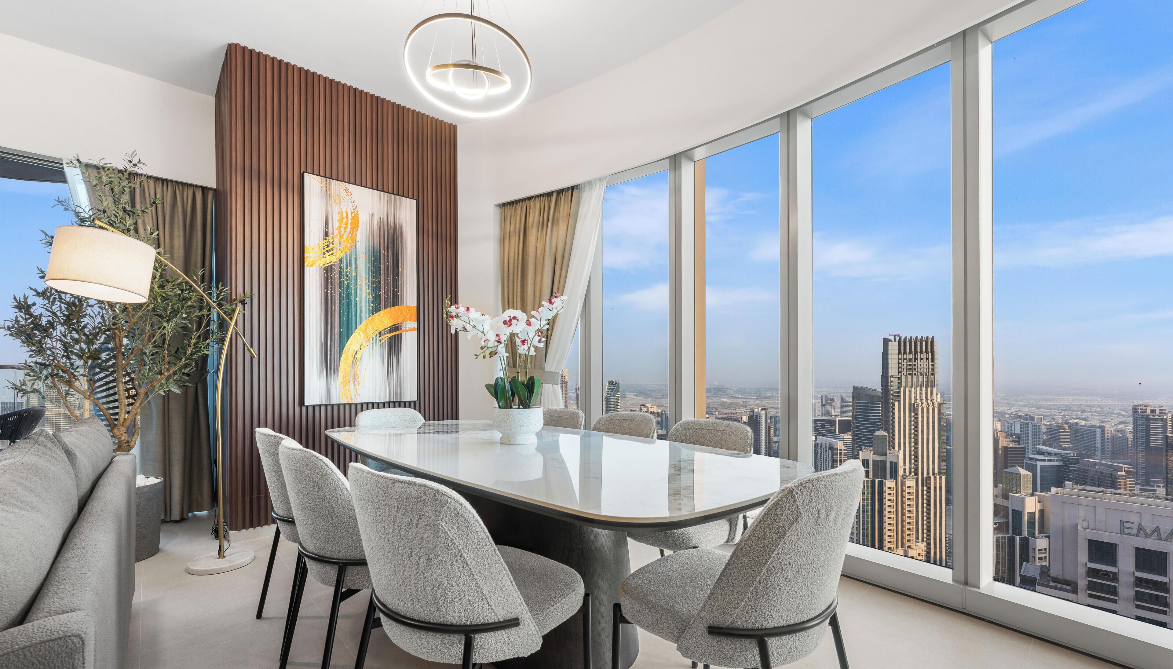 A modern dining area with a glass table and chairs, a large abstract painting, and a view of the cityscape through large windows.