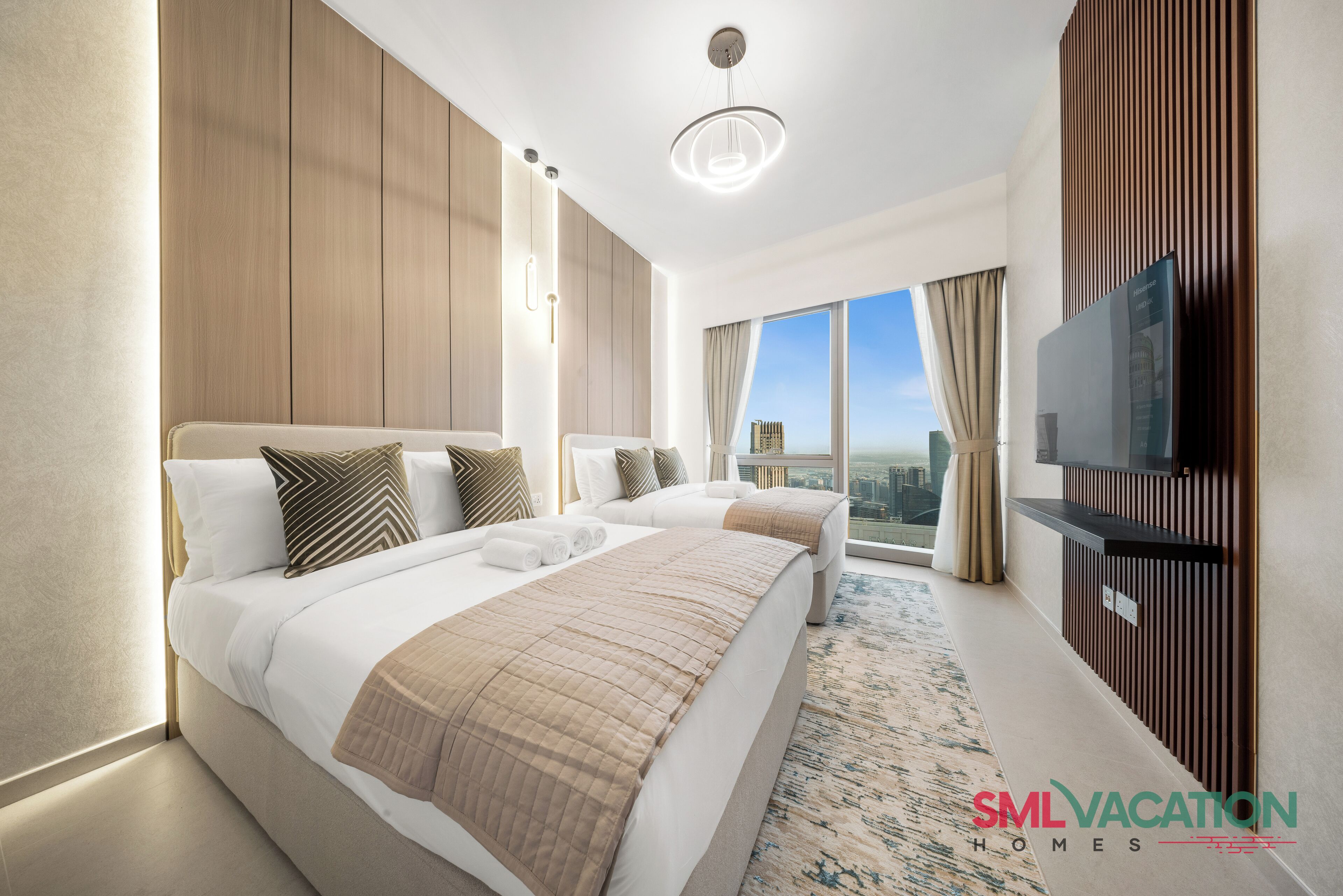 A modern hotel room with two beds, a large window with a city view, a flat-screen TV, and a wooden accent wall.