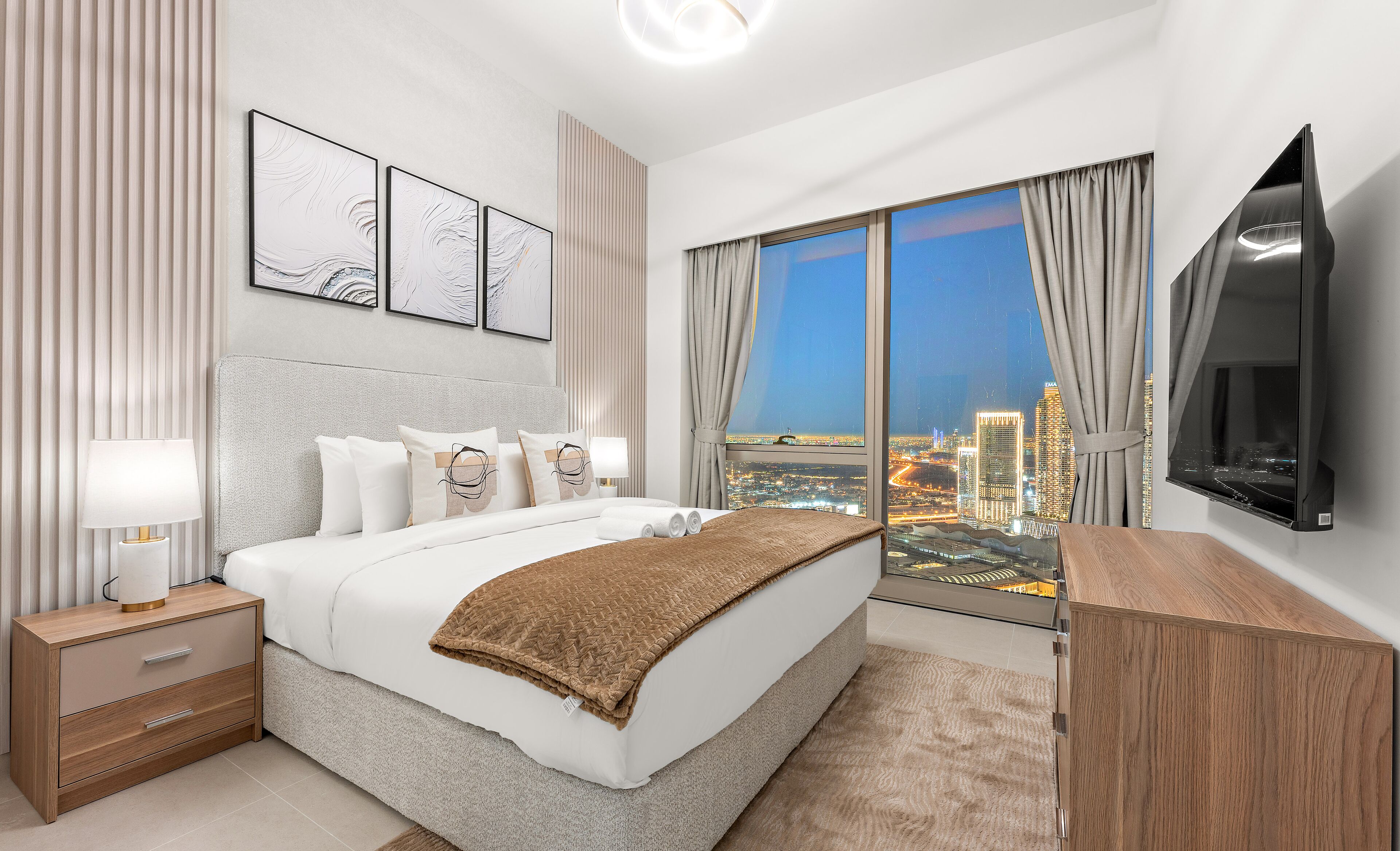 A modern hotel room with a large bed, a bedside table with a lamp, a TV mounted on the wall, and a city view through large windows.