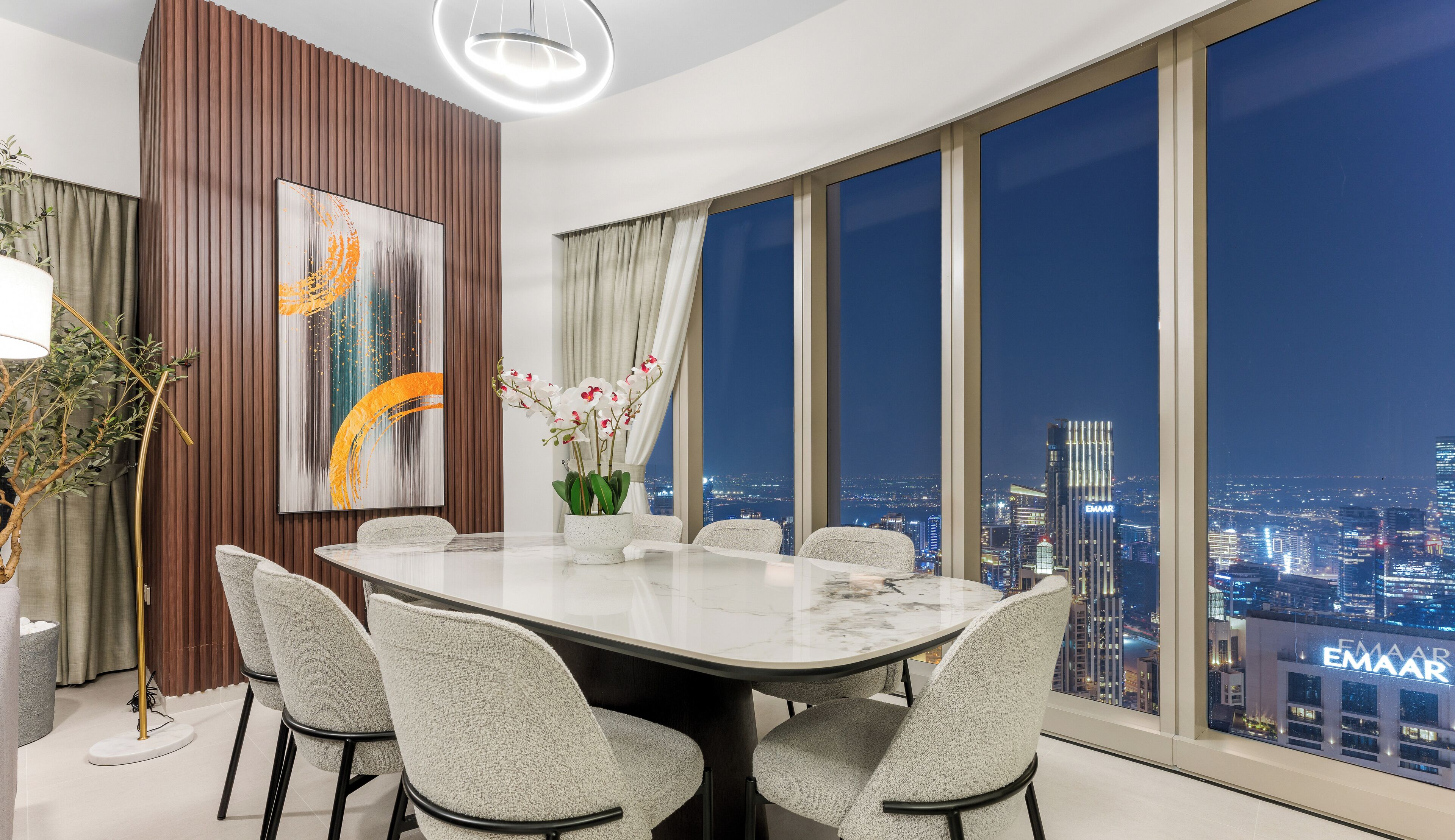 A modern dining area with a large table and chairs, a city view through large windows, and a decorative painting on the wall.