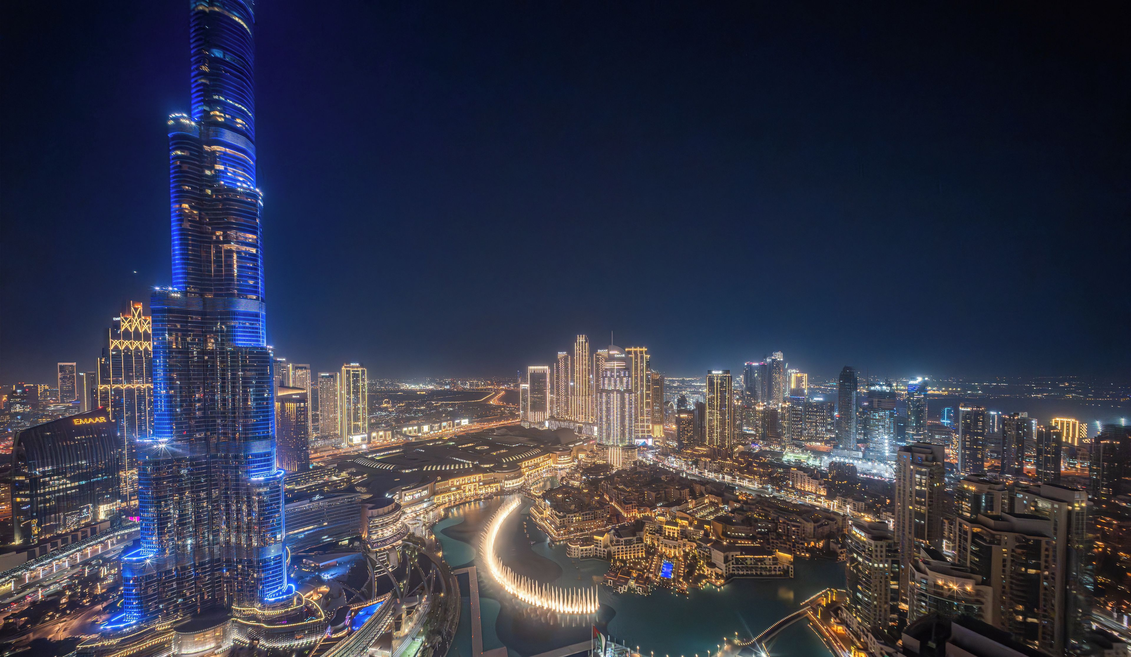 A nighttime cityscape with a prominent illuminated skyscraper, a waterfront area, and a dense cluster of surrounding buildings.