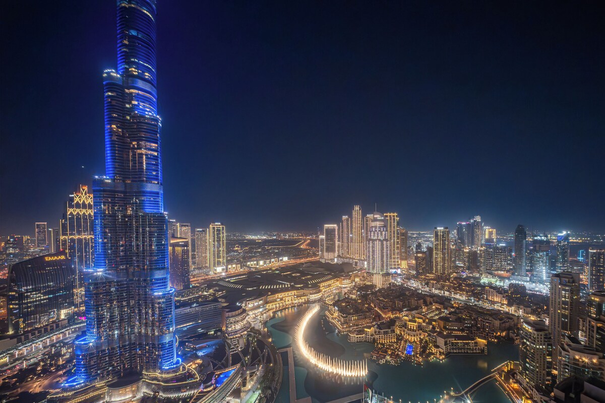 A nighttime cityscape with a prominent illuminated skyscraper, a waterfront area, and a dense cluster of surrounding buildings.