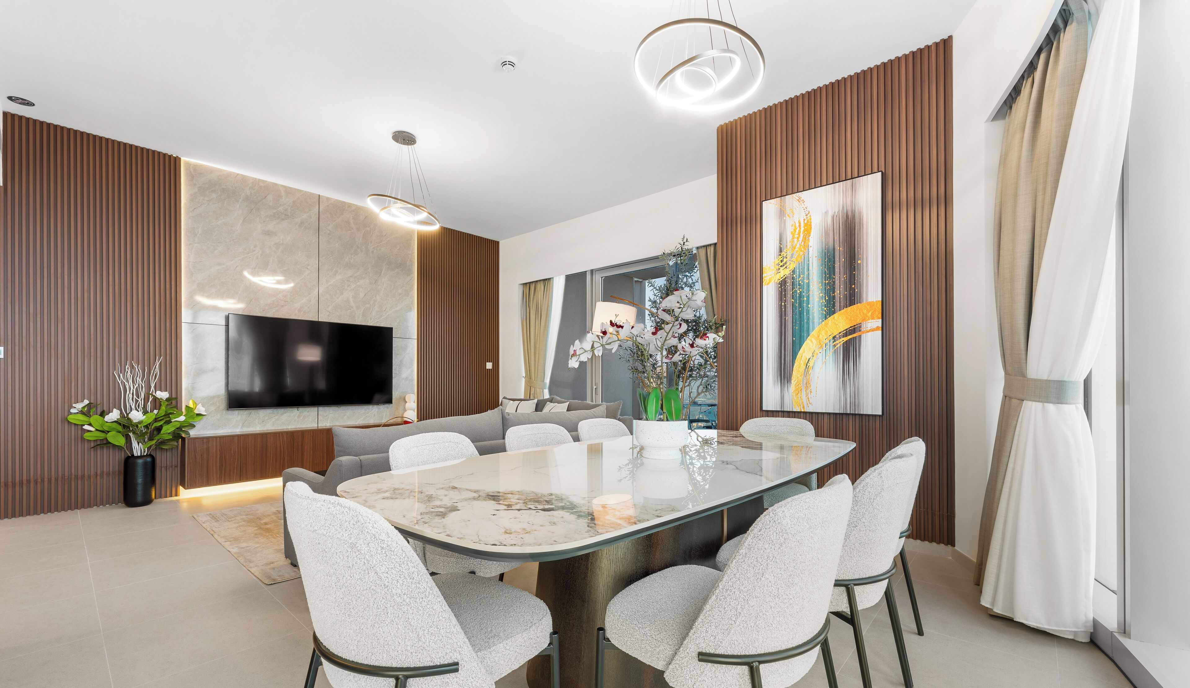A modern dining area with a marble table, white upholstered chairs, a flat-screen TV, and a large abstract painting on the wall.