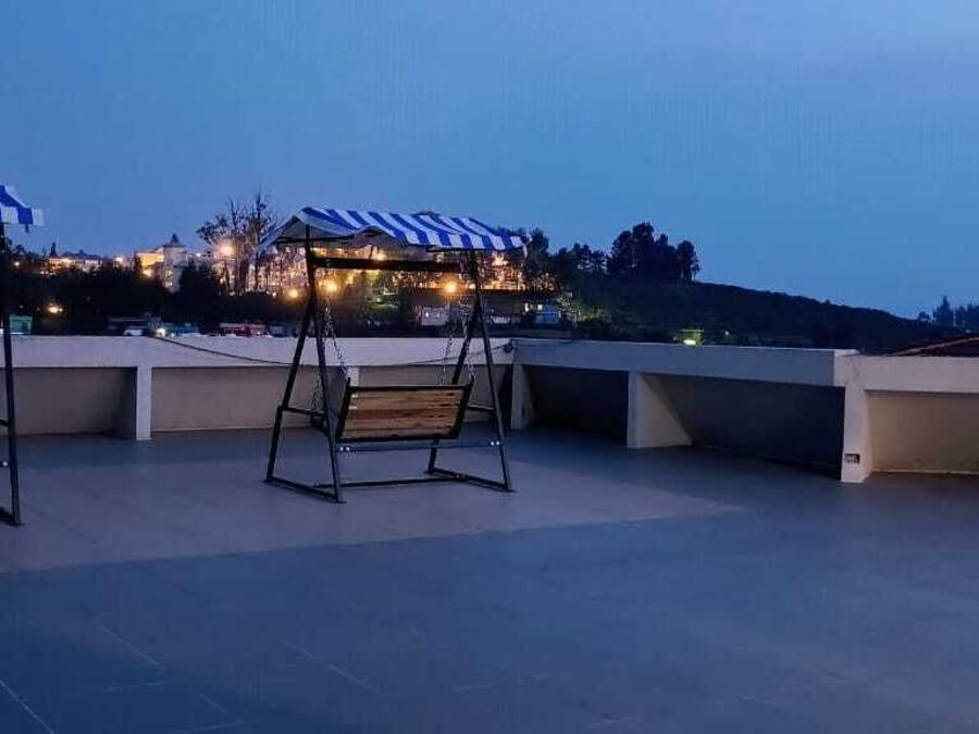 Terraza en la azotea con dos asientos tipo columpio y una zona de estar cubierta, con vistas a un edificio iluminado y un paisaje montañoso.