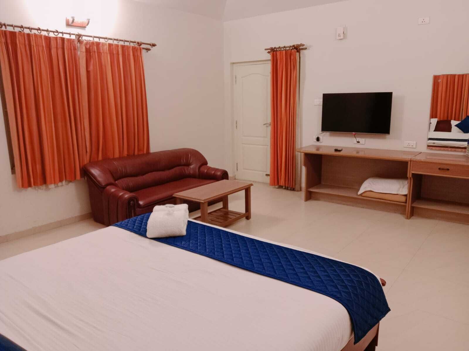 A hotel room with a bed, a brown leather sofa, a wooden desk with a TV, and orange curtains.