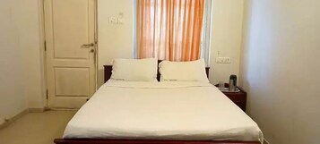 A single bed with white bedding in a room with a closed door and a window with orange curtains.