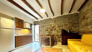 A kitchen with a stone wall, wooden beams, a yellow sofa, and a dining area. - " Village Center Apartments - Apt 1 - 2 " (Bari Sardo)