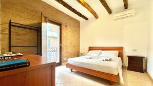 A bedroom with a wooden bed, a bedside table with books, a window with a view, and a tiled floor. - " Village Center Apartments - Apt 1 - 2 " (Bari Sardo)