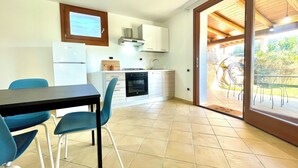 Een kleine keuken met een koelkast, fornuis en kastjes. Er is een eethoek met een tafel en stoelen.