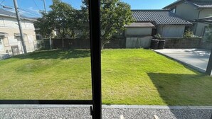 Une vue à travers une fenêtre donnant sur un quartier résidentiel, avec une cour verdoyante, un banc en bois et un bâtiment doté d’un toit en tuiles.
