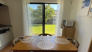 Un coin repas avec une table dressée pour quatre personnes, une cuisine en arrière-plan et une vue sur un jardin par une porte coulissante.