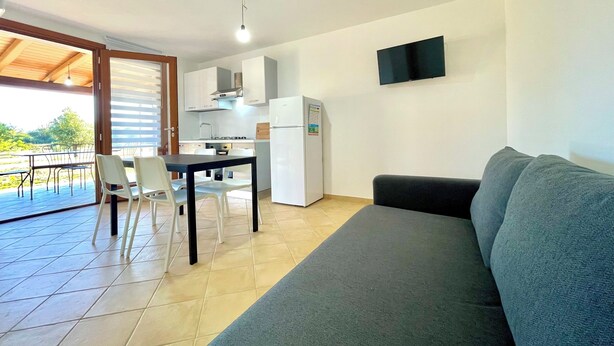 A compact living space with a kitchenette, a dining area with a table and chairs, a sofa, and a TV mounted on the wall. - " Cronta Apartments with patio " (Bari Sardo)