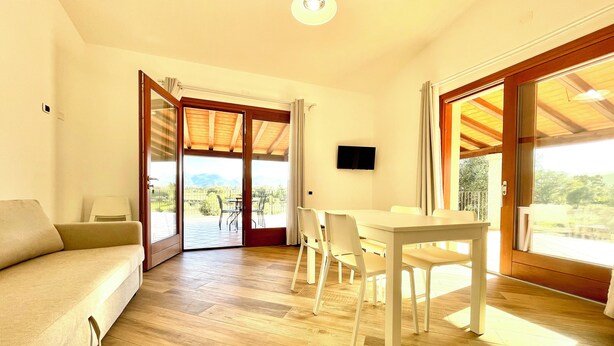 A room with a sofa, a dining table with chairs, a TV, and large sliding glass doors leading to an outdoor area with a view of the mountains. - " Cronta Apartments Deluxe with Terrace " (Bari Sardo)