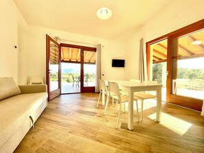 A room with a sofa, a dining table with chairs, a TV, and large sliding glass doors leading to an outdoor area with a view of the mountains. - " Cronta Apartments Deluxe with Terrace " (Bari Sardo)