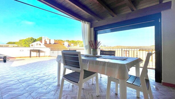 A covered outdoor patio with a table and chairs, a view of buildings in the distance, and a tiled floor. - " Beach House with Terrace " (Bari Sardo)