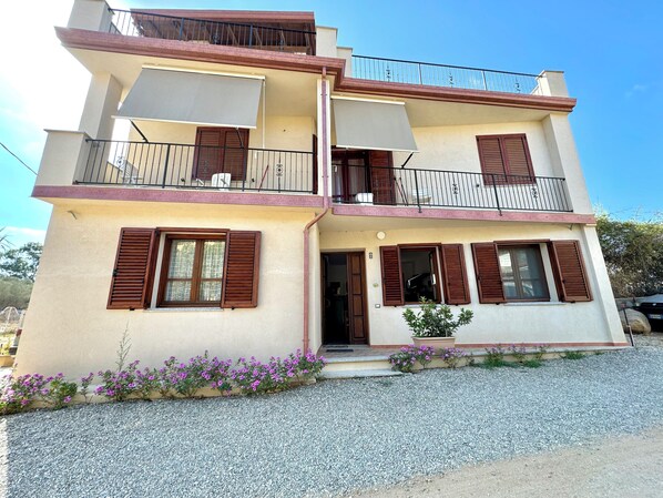 A two-story building with balconies, a gravel driveway, and a small garden with purple flowers.