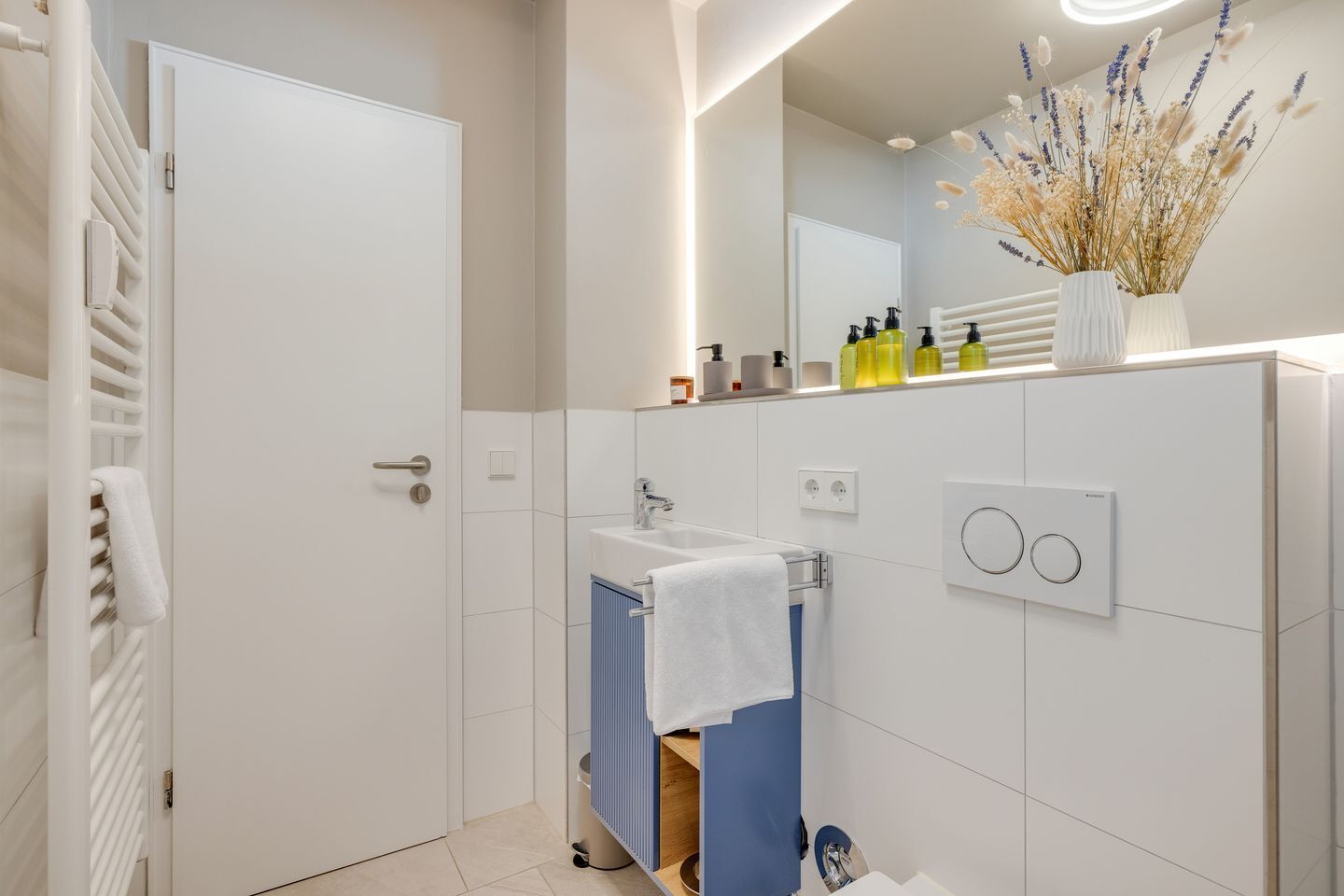 A modern bathroom with a white door, a sink with a towel rack, and a mirror with a decorative vase.