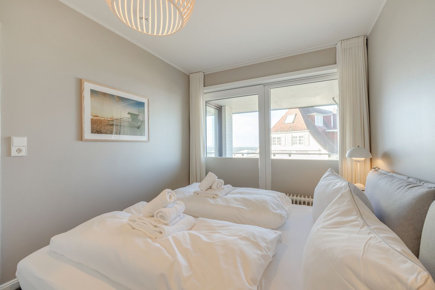 A hotel room with a bed, a window with curtains, a lamp, and a painting on the wall.