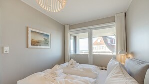 A hotel room with a bed, a window with curtains, a lamp, and a painting on the wall. - Strandperle - "Strandperle on Sylt: Your dream vacation with sea views and comfort!" (Wenningstedt-Braderup (Sylt))