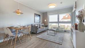 A modern living room with a dining area, large windows, and a view of a building. - Strandperle - "Strandperle on Sylt: Your dream vacation with sea views and comfort!" (Wenningstedt-Braderup (Sylt))