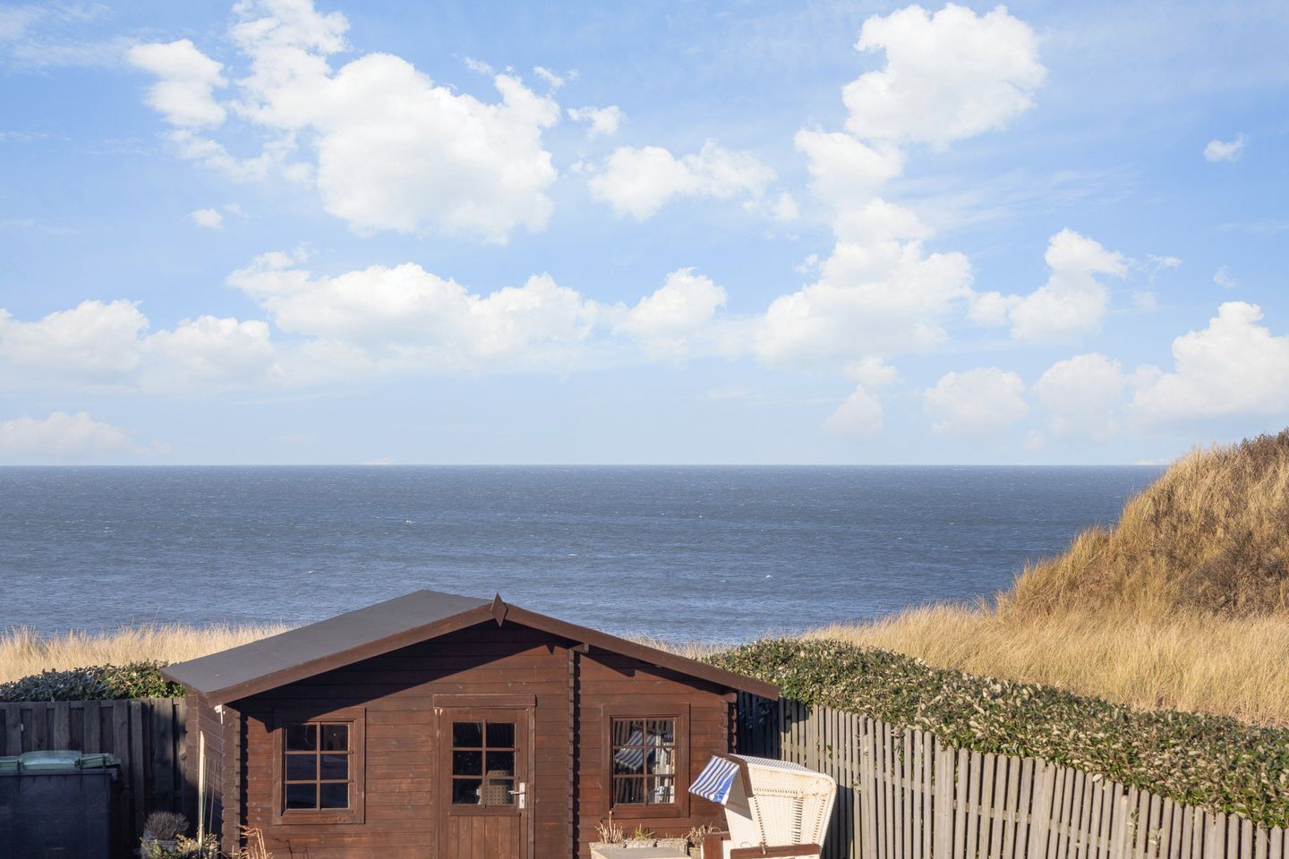A wooden cabin with a view of the sea, a fenced garden, and a chair.