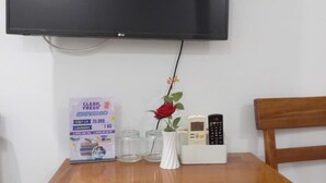 Egy szoba, melyben a falra van szerelve egy síkképernyős televízió, egy fehér vázában piros rózsa áll egy fa asztalon, emellett van egy kis hűtőszekrény, és a távirányító az asztalon található.