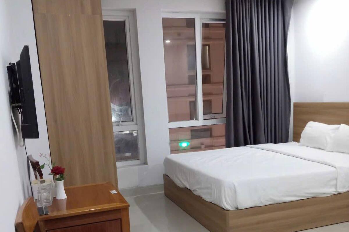 A hotel room with a bed, a wooden desk, a chair, a TV mounted on the wall, and a window with curtains.