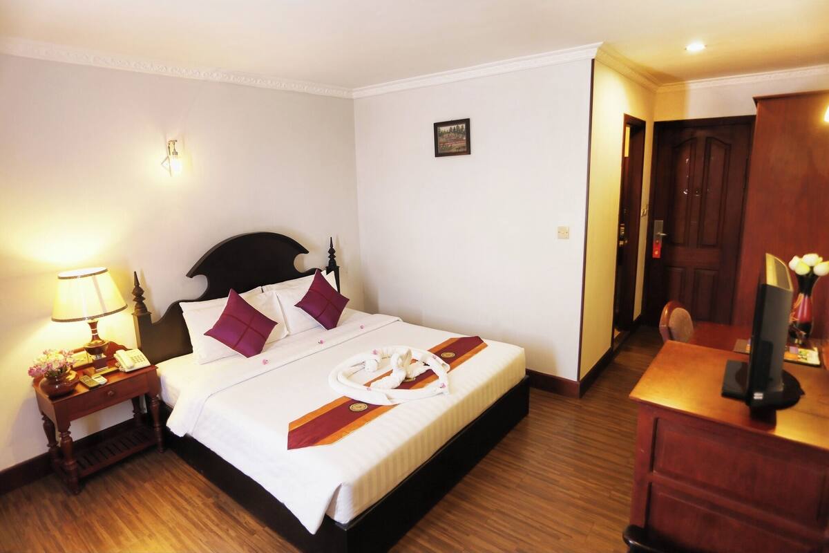 A hotel room with a neatly made bed, a bedside table with a lamp, a desk with a computer, and a door leading to another room.