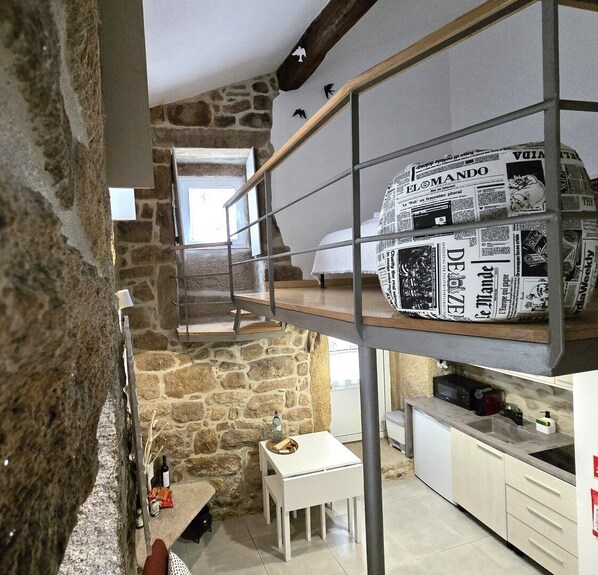 A rustic room with a stone wall, a wooden staircase, a white bed, and a kitchen area with a newspaper-covered shelf.