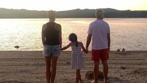 A family of three standing on a sandy beach facing a calm body of water with hills in the background.