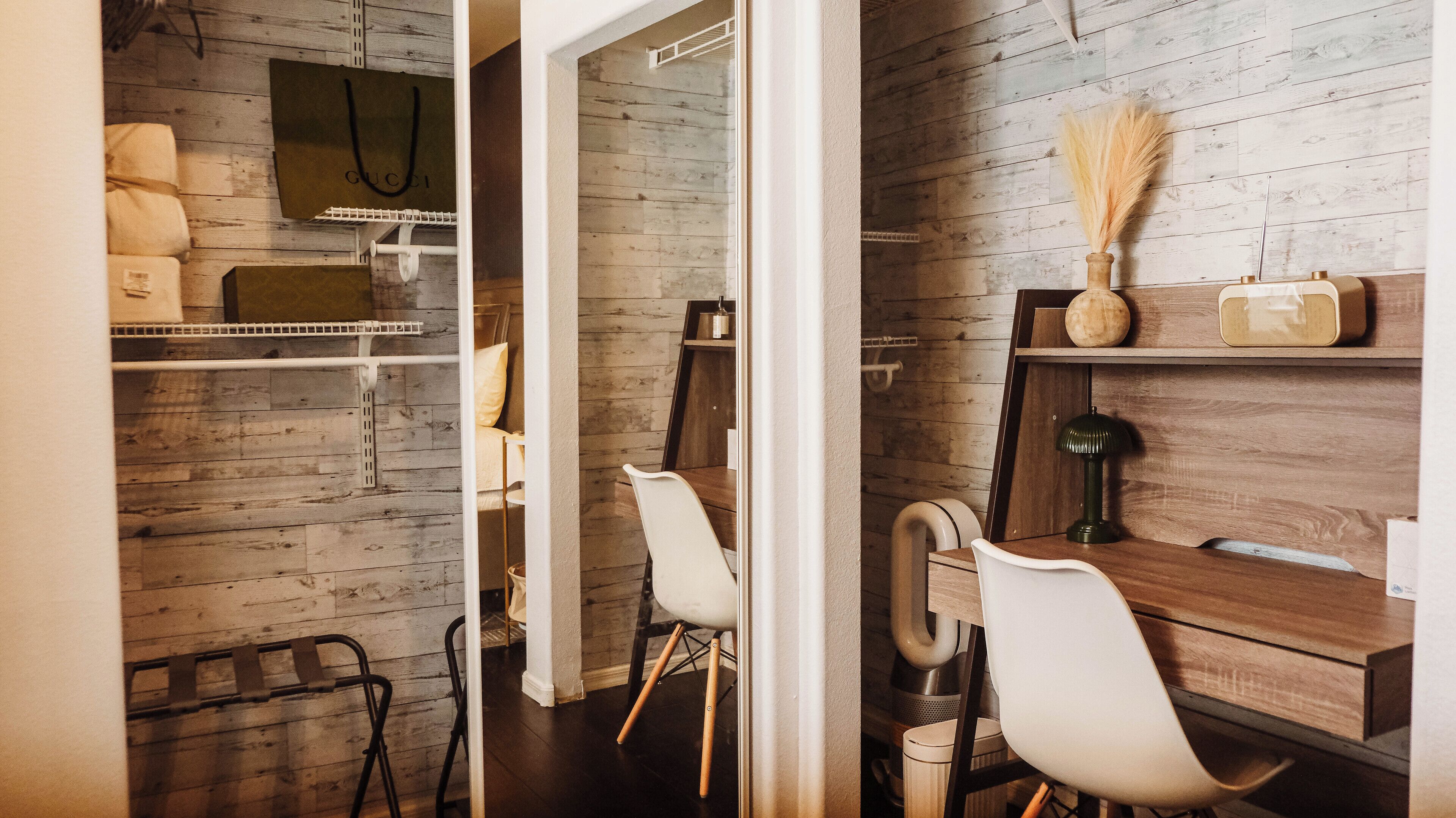 A room with a wooden wall, a white chair, a wooden desk, and a small shelf with a vase and a lamp.