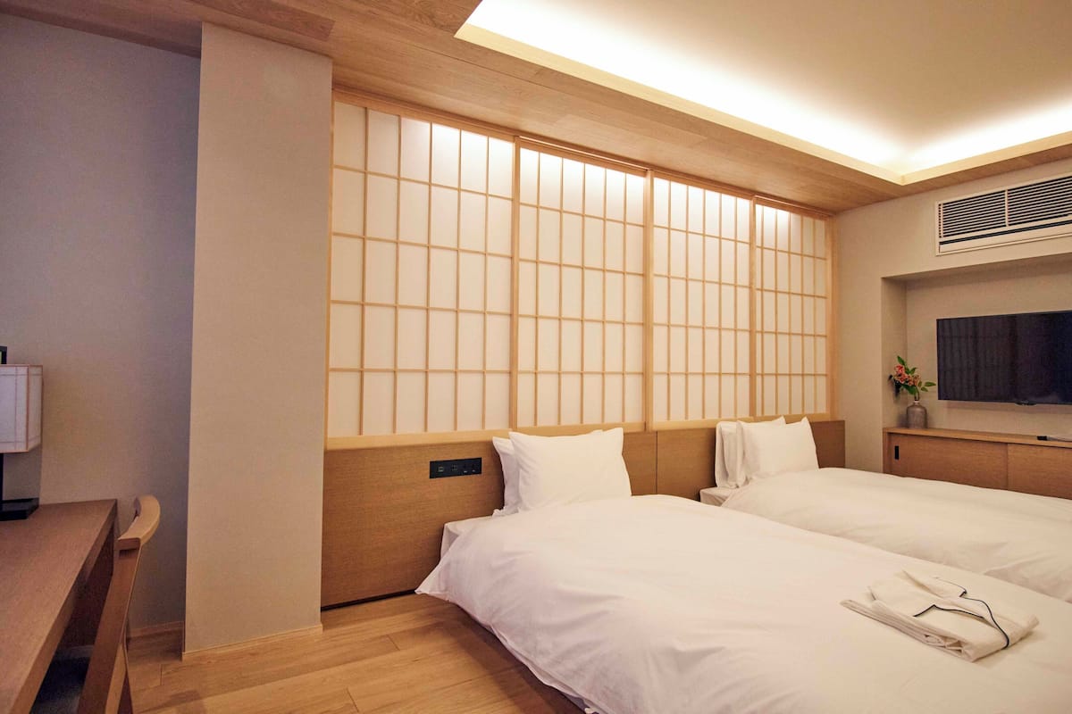 Camera d'albergo con due letti, una testiera in legno, un comodino con una lampada e un'ampia finestra con vista sulla città.