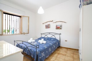 Une chambre à coucher comprenant un lit, une table de chevet, un luminaire suspendu, une fenêtre avec des barreaux et une étagère fixée au mur, ornée de deux objets décoratifs.