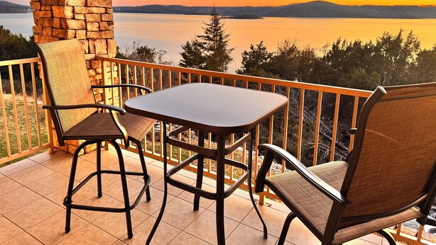 A balcony with a stone pillar, two chairs, and a table overlooking a lake at sunset.