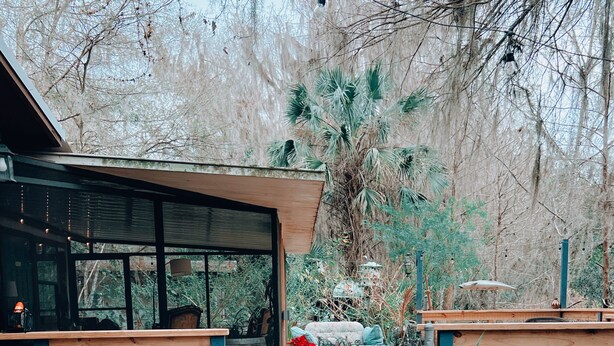 Un sendero de madera conduce a un edificio moderno con amplios ventanales, un área de descanso con mesa y sillas, y una variedad de plantas y elementos decorativos.