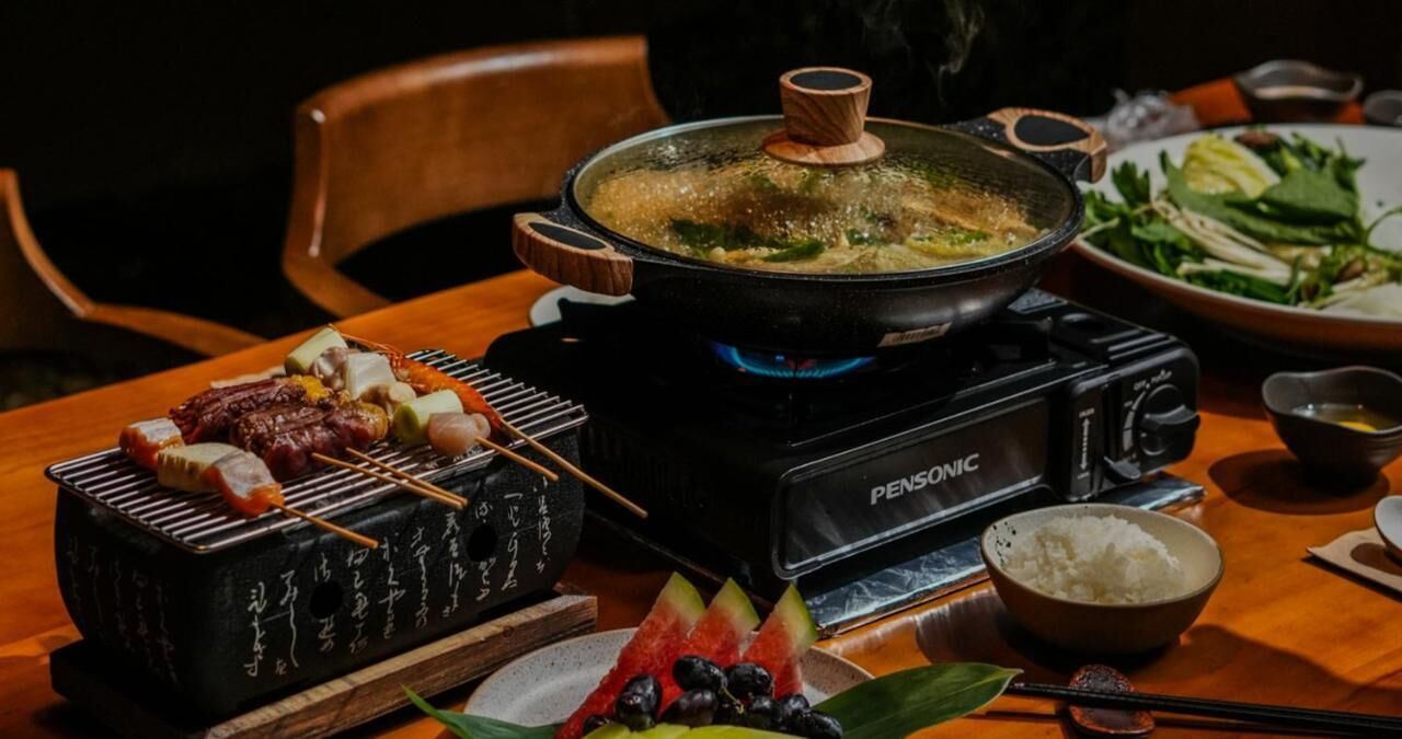 A traditional Japanese hot pot setup with a PensoNIC portable stove, skewered meats, a bowl of rice, and a side dish of vegetables.