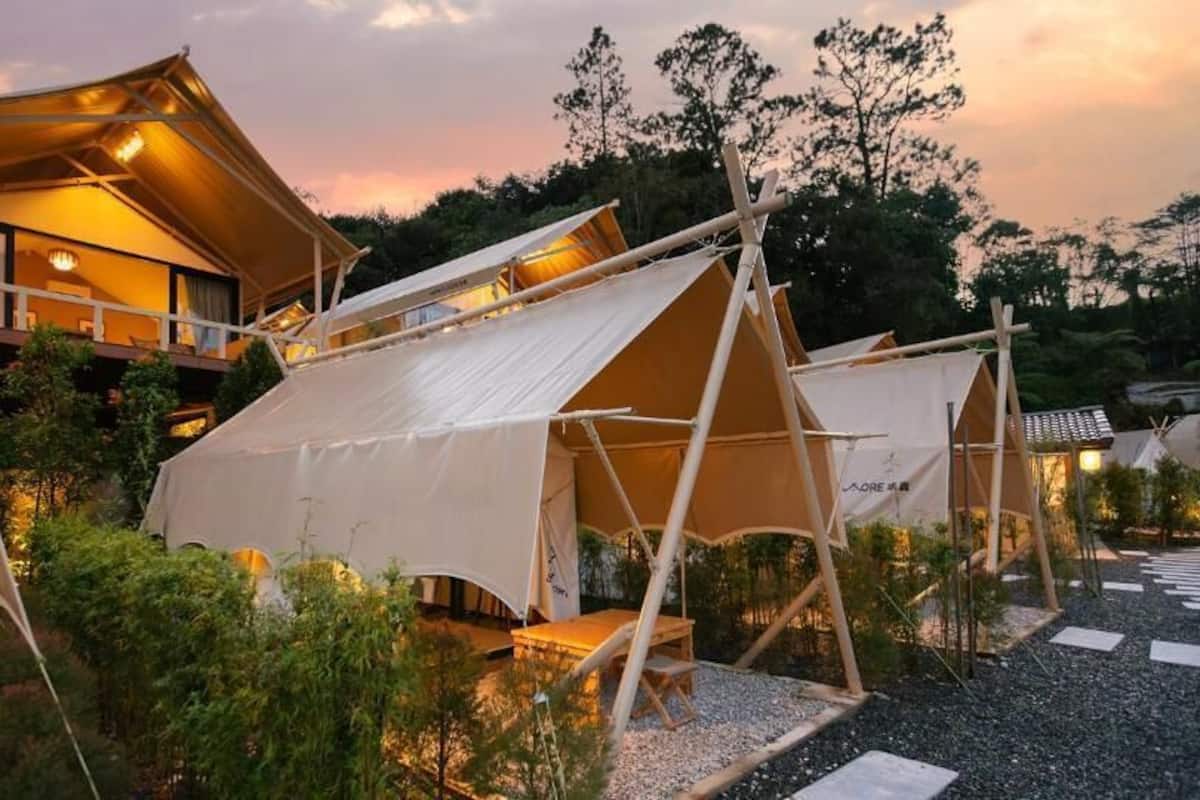 A campsite with multiple tents and a cabin with a balcony, surrounded by trees and a gravel path.
