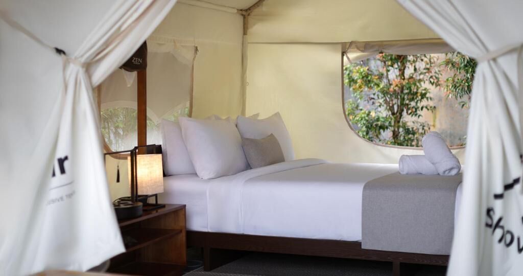 Una cama con ropa de cama y almohadas blancas, una lámpara de noche y una ventana con vistas a árboles.