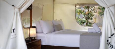Una cama con ropa de cama y almohadas blancas, una lámpara de noche y una ventana con vistas a árboles.