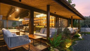 Un espacio moderno para disfrutar del aire libre con piso de madera, un sofá, una silla y una mesa. Hay una pared de vidrio con estanterías y una escalera que sube a un nivel superior.