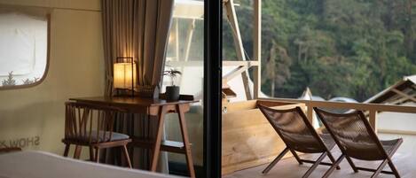 A room with a wooden desk, two wicker chairs, a lamp, and a view of a forest through a window.