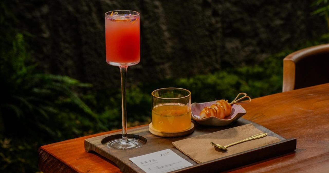 A wooden table with a menu, a tall glass of red drink, a short glass of yellow drink, and a small bowl of snacks.