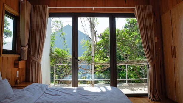 A bedroom with a large sliding glass door leading to a balcony, wooden walls, and a bed with white bedding. - La Maison Dalat (Lang Biang - Da Lat)