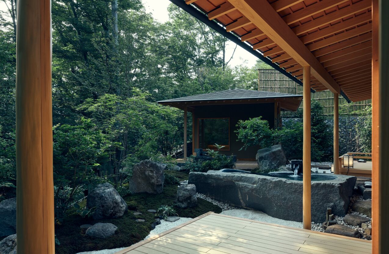 A traditional Japanese garden with a wooden pavilion, rock formations, and a small water feature.