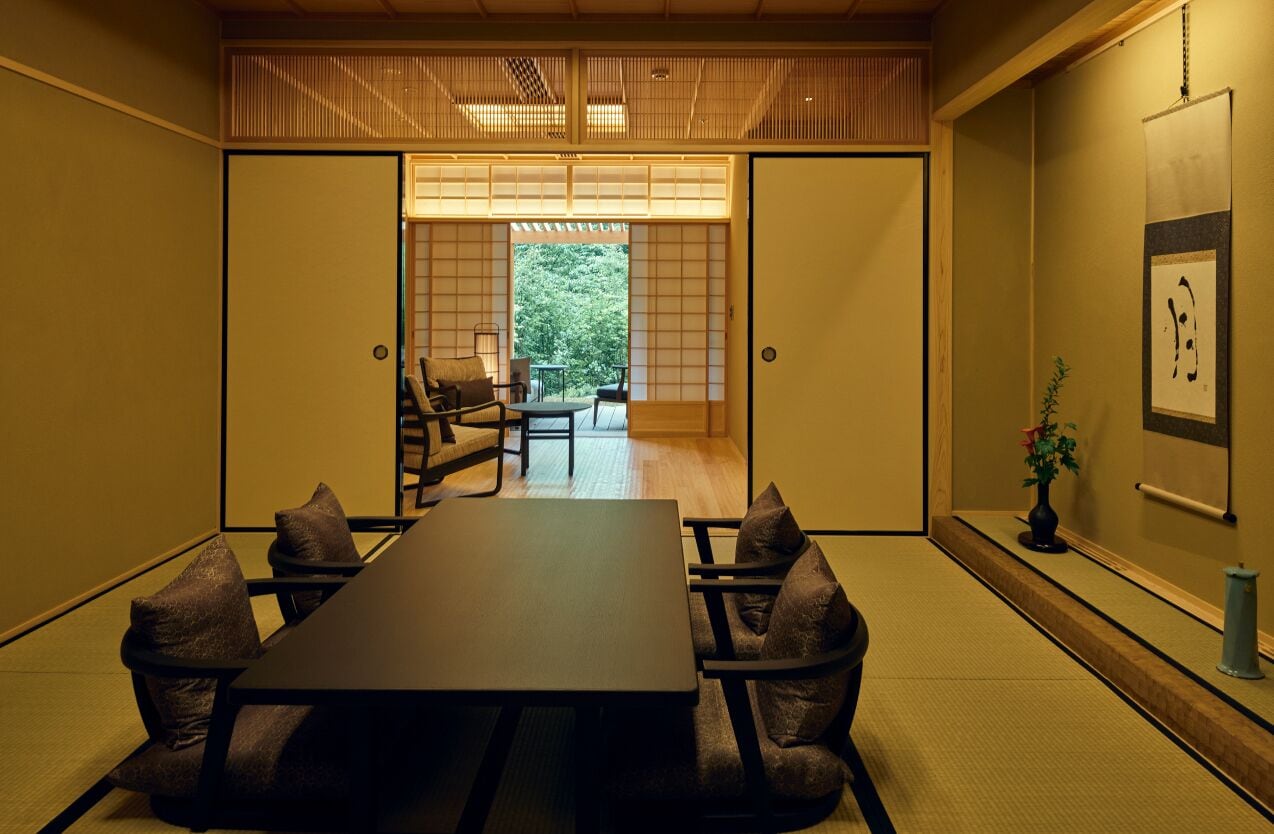 Um quarto tradicional japonês com chão de tatame, uma mesa baixa com almofadas e portas corrediças que dão para um jardim.