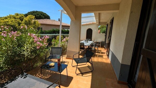 An outdoor patio with a table and chairs, surrounded by greenery and a building in the background.